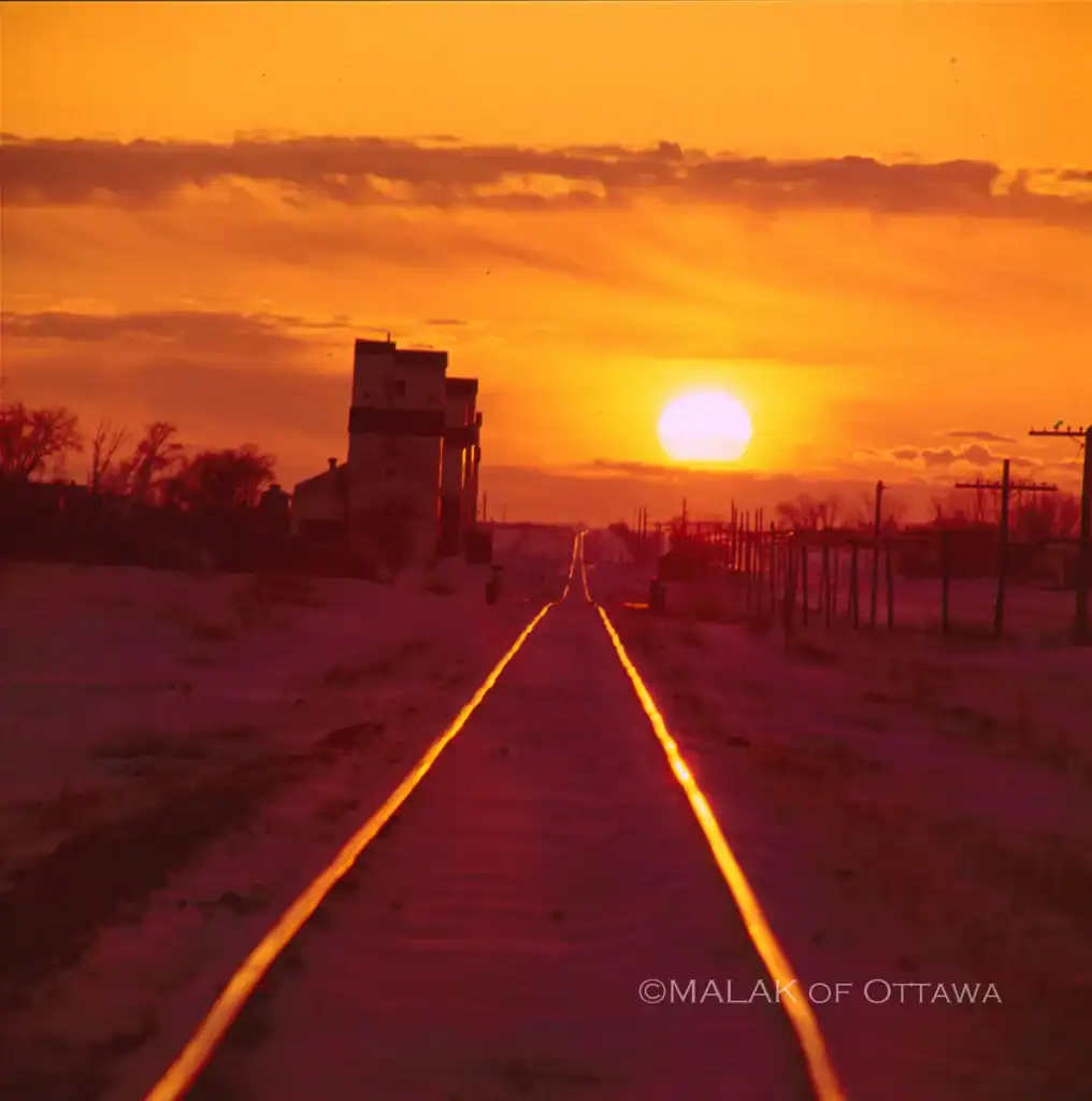 Sunset over railway tracks in Ottawa with vibrant orange sky and silhouettes of buildings and trees.
