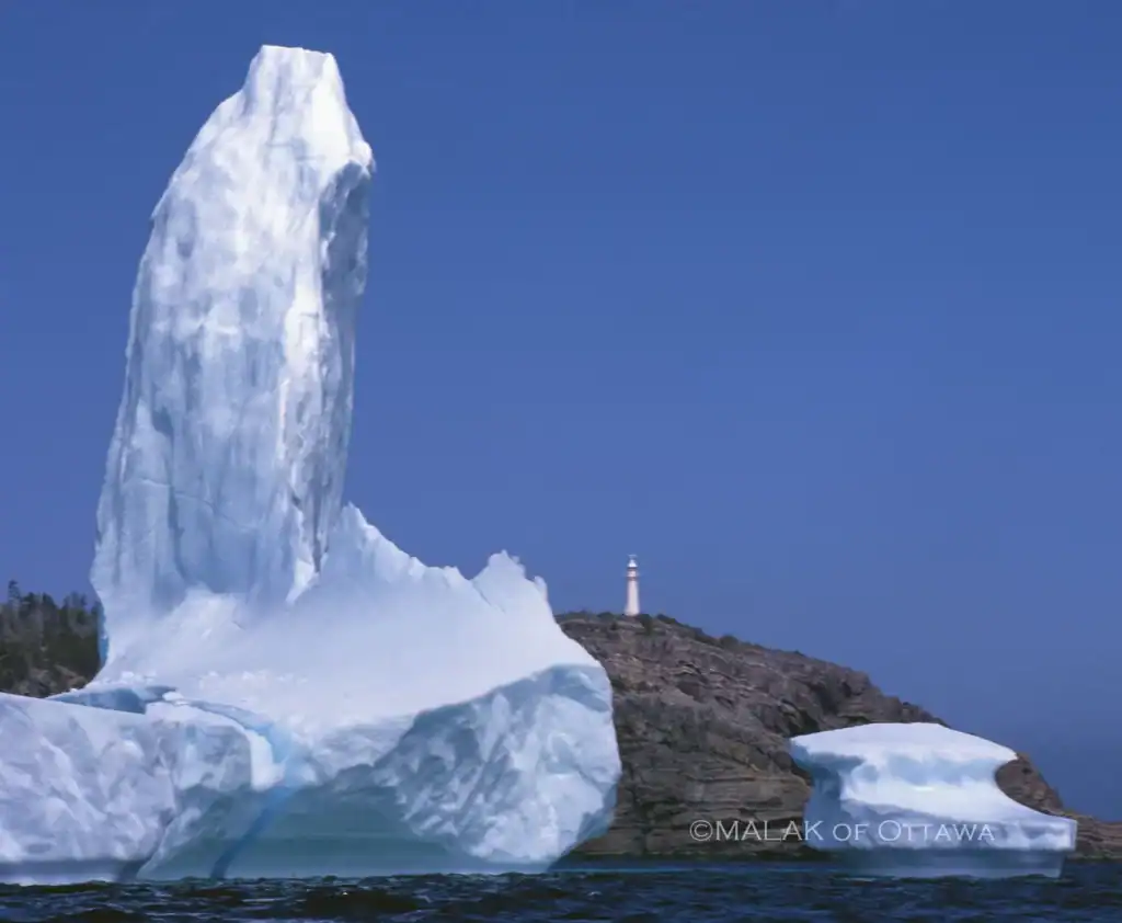 Iceberg with towering ice formation and smaller chunks floating nearby, against a clear blue sky.