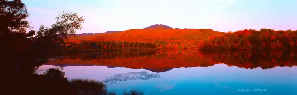 Scenic view of a calm lake with autumn-colored trees and distant hills at sunset.