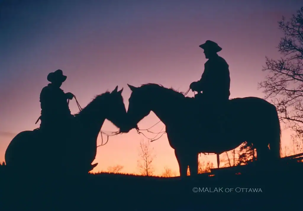 Silhouettes of two horseback riders at sunset with a colorful sky in the background.