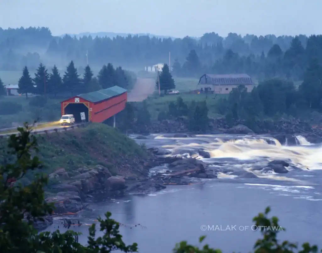 Scenic view of a river with waterfalls, farm buildings, and a red barn in a lush, green landscape ne.